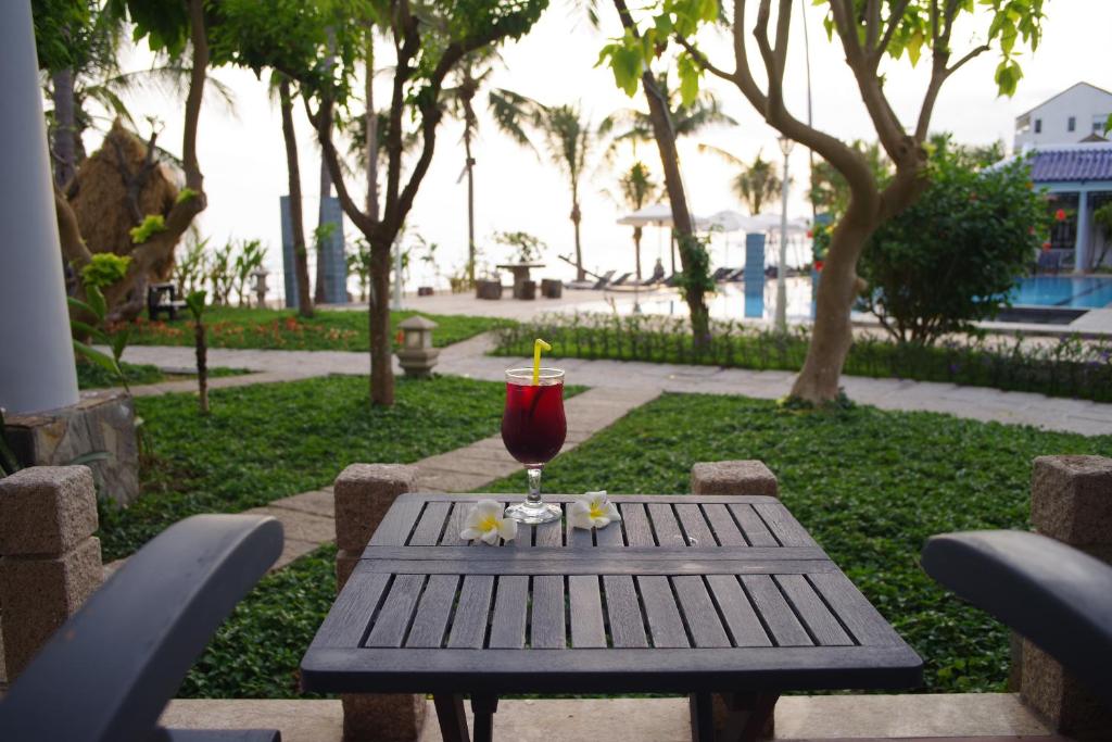 Grezna terase ar galdu, uz kuras atrodas kokteilis ar salmiņu un ziedu, apkārt zāle un palmām. Fons veido mierīgu ainavu ar baseinu un jūru.