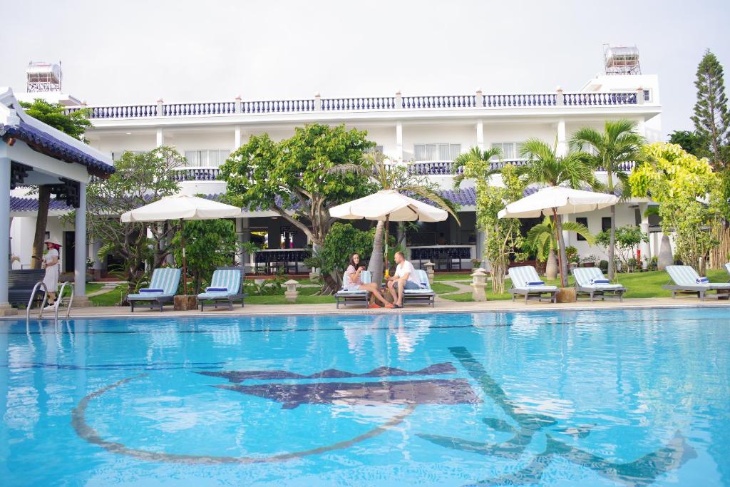 Peldbaseins ar saulessargiem un atpūtas krēsliem, apkārtne ar zaļumiem un viesnīcas ēka fonā.