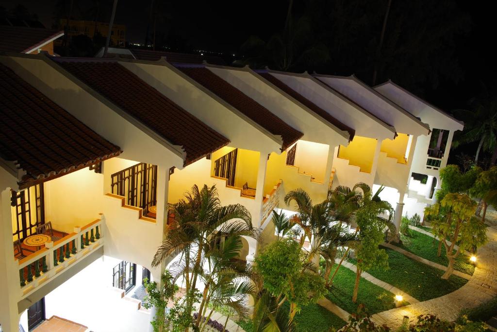 Nakts ainava ar baltiem māju jumtiem un zaļiem krūmiem, izgaismota ar dārza lampām. Mājas veido patīkamu atmosfēru, kas aicina uz atpūtu.