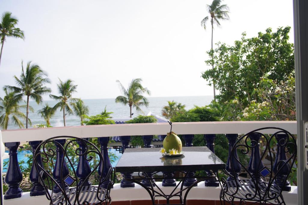 Skats uz jūru no balkona ar melnu galdu un divām dzelzs krēsliem, uz galda novietota kokosrieksts ar ziediem. Apkārt aug palmās un zaļumi.
