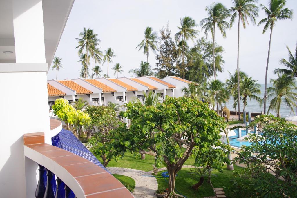 Tropu ainava ar palmu kokiem un baseinu, redzama no viesnīcas balkona. Ēka ar sarkaniem jumtiem un zaļo dārzu piekrastē.