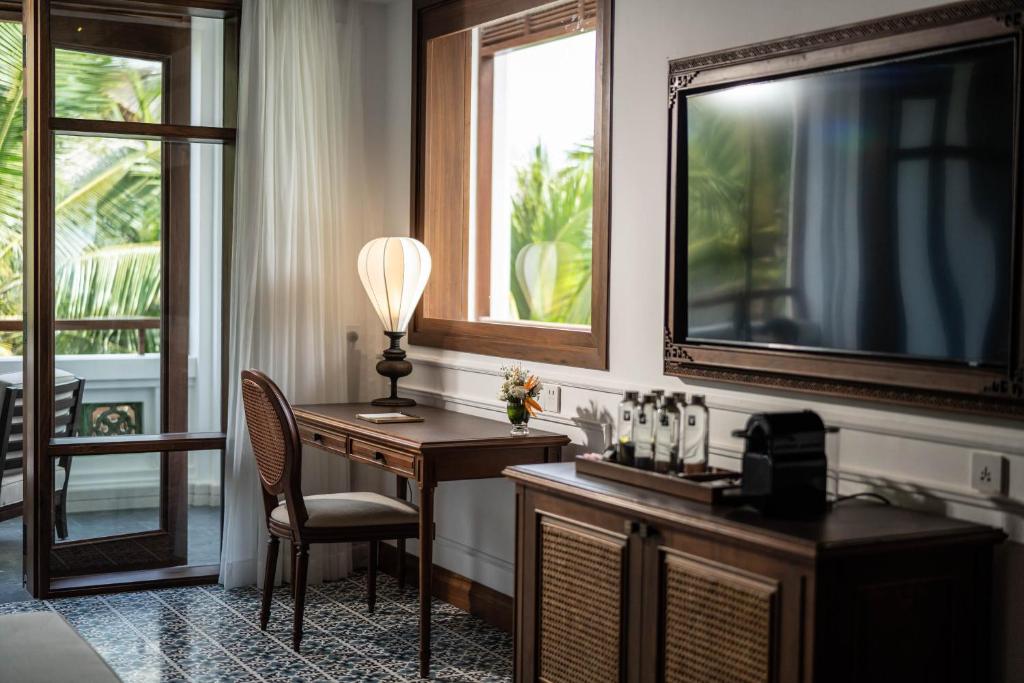 Mūsdienīgs viesnīcas numurs ar elegantu koka mēbelēm, stilīgu lampu un televizoru, kas piedāvā skatu uz zaļajiem palmām ārpus loga.