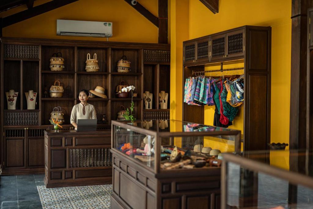 Veikals ar koka interjeru, kurā redzama sieviete pie letes ar datoru, apkārt pirkumiem, piemēram, somām un peldkostīmiem. Sienas ir dzeltenas, radot siltu atmosfēru.