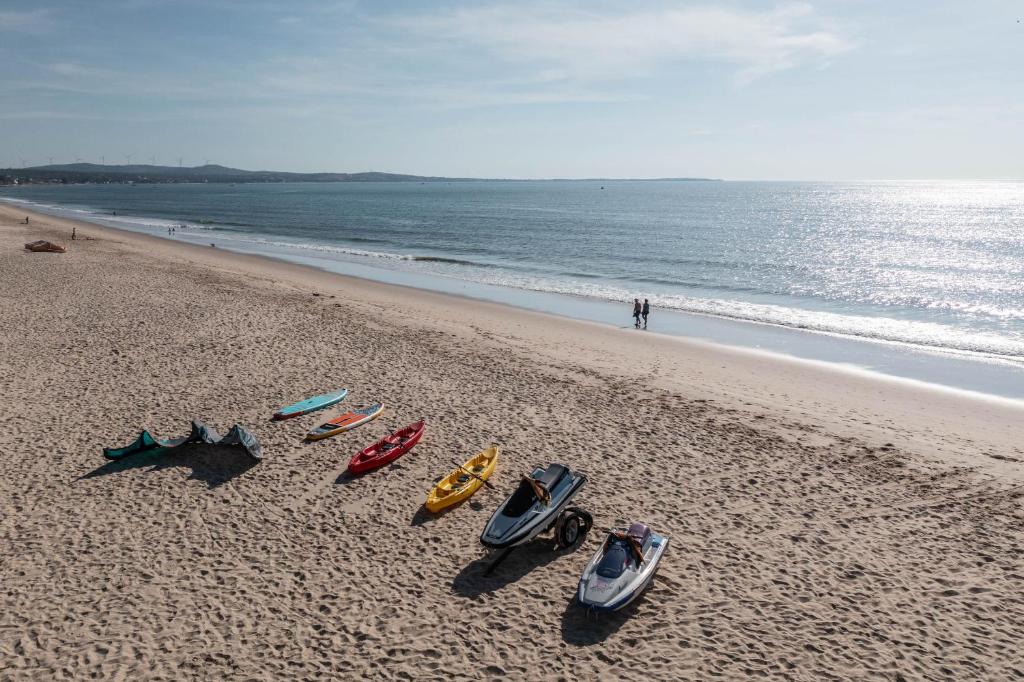 Pludmales ainava ar dažādām laivām, kas novietotas smiltīs, un cilvēkiem, kas pastaigājas gar krastu. Jūra ir mierīga un saulaina diena.