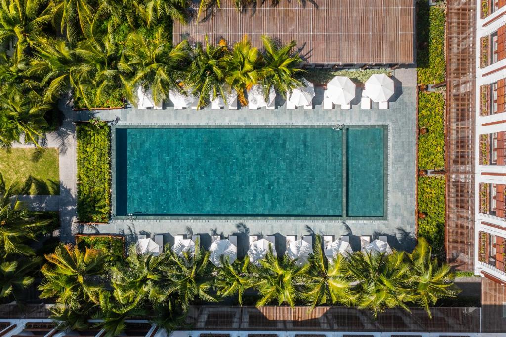 Lidojuma skats uz modernu peldbaseinu, ko ieskauj zaļi palmu koki un atpūtas krēsli ar saulessargiem. Baseins ir centrālais elements relaksējošā atpūtas zonā.