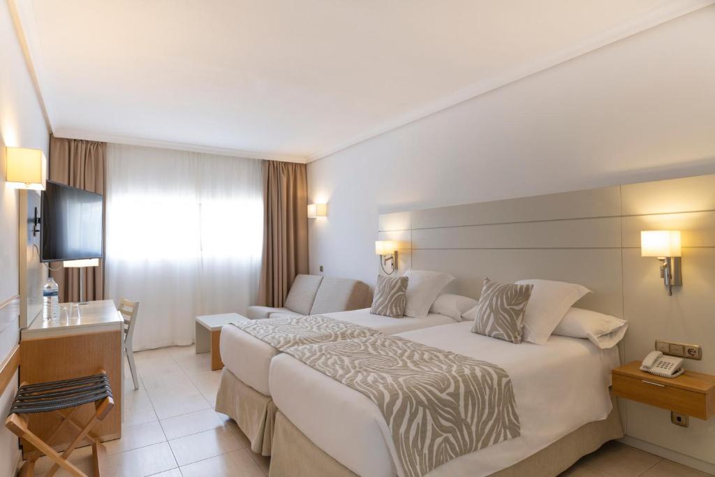 Mūsdienīgs viesnīcas numurs ar divām gultām, TV, dīvānu un galdu, kas apgaismots ar dabīgu gaismu. Interjers ir gaišs un elegants, radot relaksējošu atmosfēru.