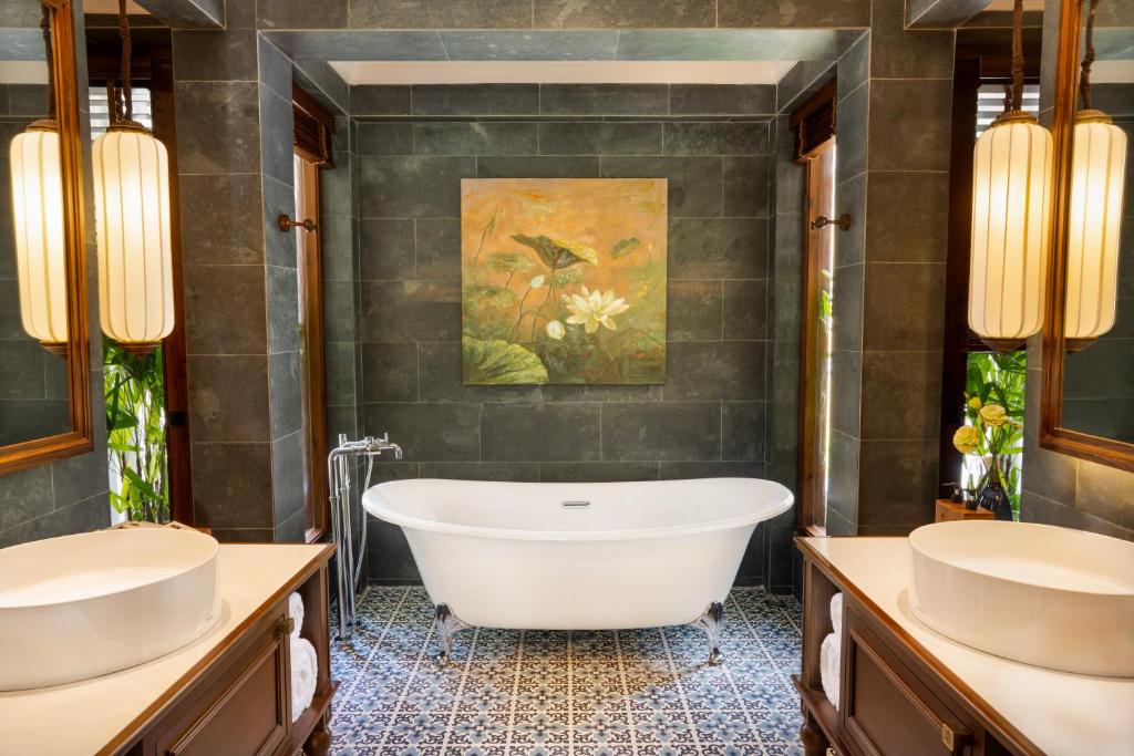 Elegants vannas istabas interjers ar brīvi stāvošu vannu, mūsdienīgām lampām un gleznām uz sienām, radot relaksācijas atmosfēru. Stikla flīzes un koka akcenti piešķir telpai siltumu un stilu.