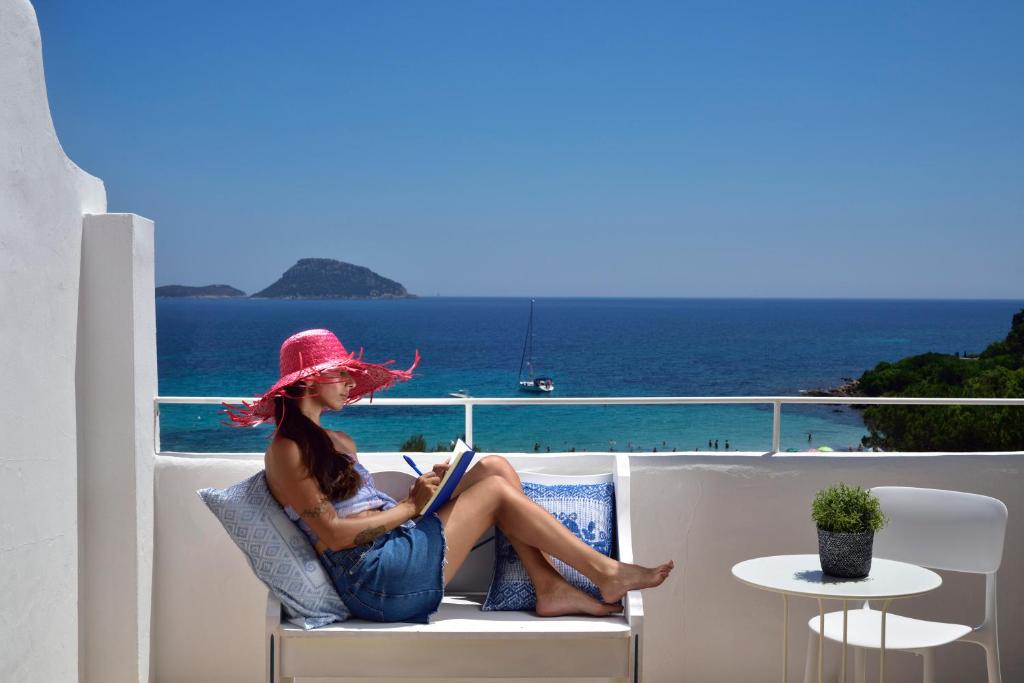 Sieviete ar rozā cepuri relaksējas uz balkona, lasot grāmatu, kamēr fona ir skaista jūras ainava un saulaina debess.
