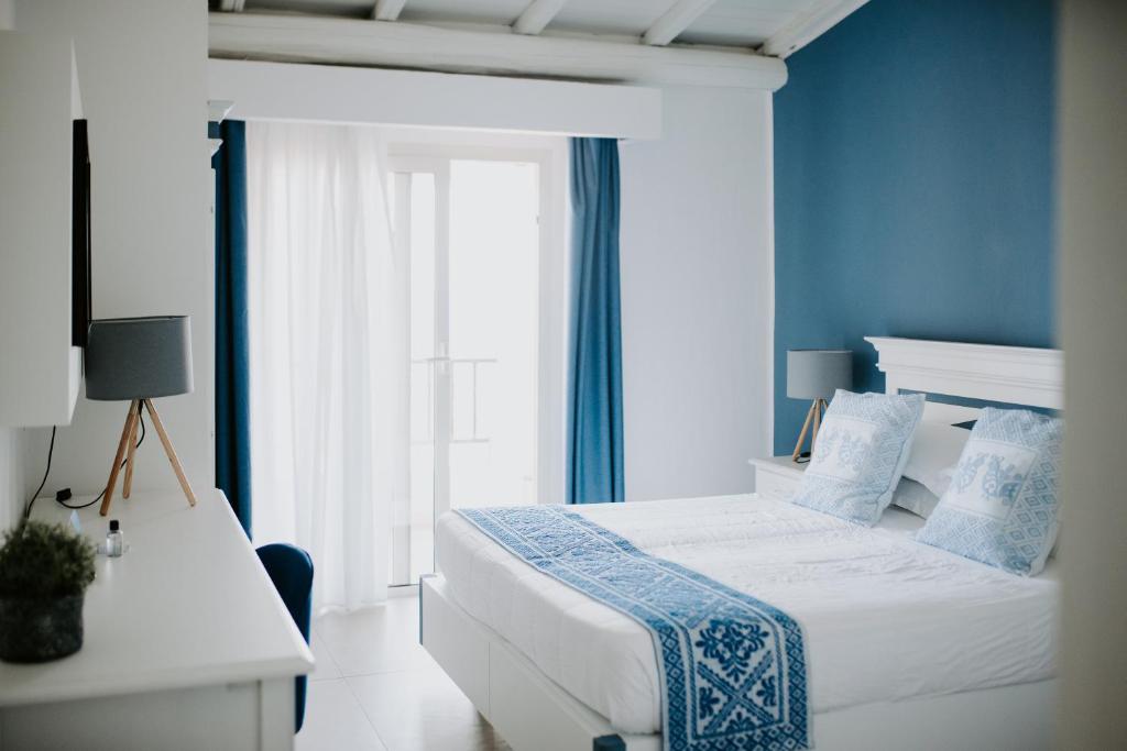 Mūsdienīgs guļamistabas interjers ar baltiem un ziliem akcentiem, iekārtots ar ērtu gultu un televizoru. Logi ar caurspīdīgām aizkariem ļauj ienākt dabīgai gaismai.