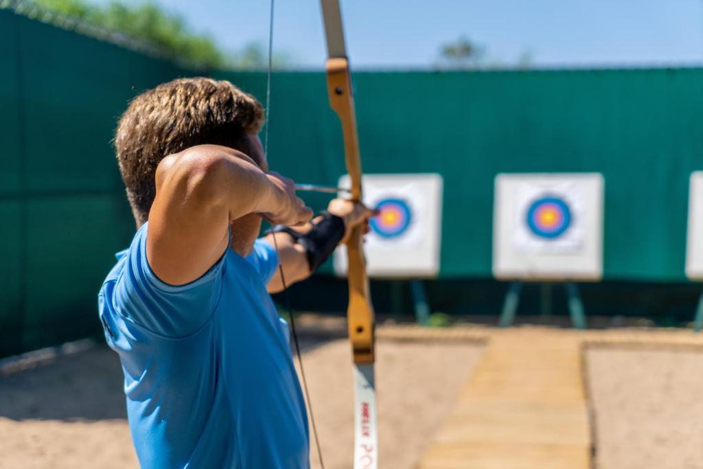Arbaleta šaušana sporta laukumā, fokuss uz šāvēju, kurš sagatavo šāvienu mērķa virzienā. Mērķi redzami fona daļā.