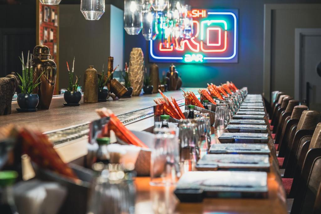 Mūsdienīgs sushi bārs ar ilgu galdu, dekorētu ar ziediem, traukiem un ēdieniem. Sarkanā un zilā neona zīme pie sienas piešķir dzīvīgumu atmosfērai.