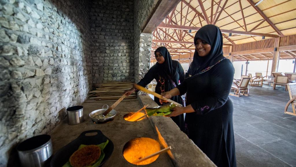 Divas sievietes gatavo ēdienu tradicionālā virtuvē, izmantojot akmens krāsni. Interjerā ir redzamas dabīgas materiālu krāsas un koka konstrukcijas.