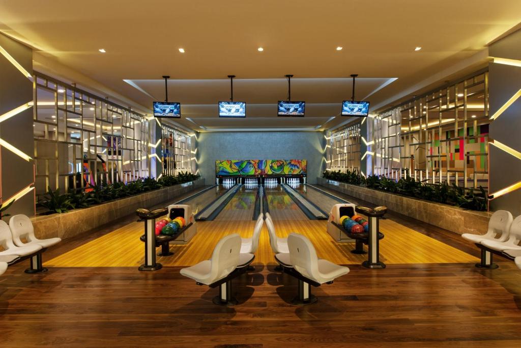 Mūsdienīgs bowlinga centrs ar spilgtu interjeru, koka grīdu un krāsainām bumbiņām, kas gaida spēlētājus. Ekrāni virs sliežu ceļa rāda spēles rezultātus.