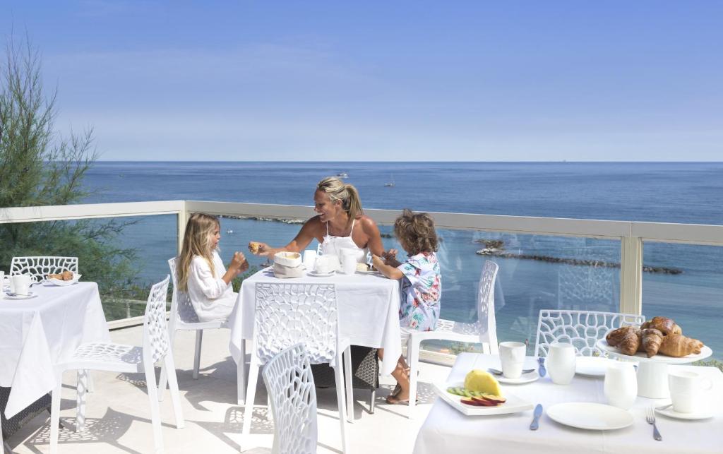 Brokastis pie jūras ar ģimeni, kur mamma un divi bērni baudīja maltīti ar skatu uz ūdeni. Uz galda ir kafija, augļi un kruasāni.