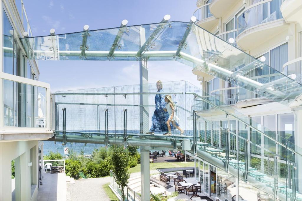 Stikla tilts pār modernas arhitektūras ēku, kas savieno divas zonas, ar skatu uz jūru. Gājēji pārvietojas pa tiltu, kas izceļas ar unikālu un stilīgu dizainu.