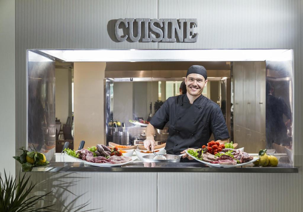 Šefpavārs, kas smaida un gatavo ēdienus, stāv pie virtuves letes, kurā redzami dažādi gaļas un dārzeņu ēdieni. Fons ir moderna virtuve ar spoguļiem un aprīkojumu.