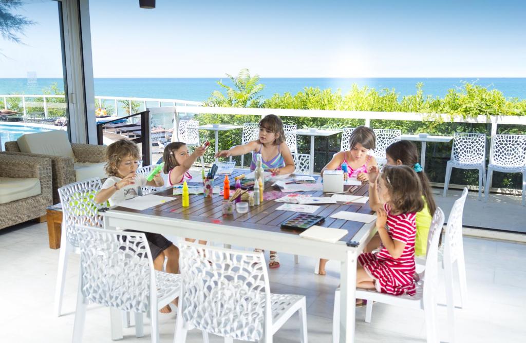 Bērni radoši darbojas pie galda, krāsojot un zīmējot, kamēr fonā redzams jūras skats. Gaisīga un radoša atmosfēra.