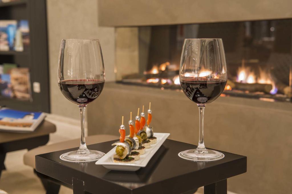 Sarkanais vīns un uzkodas tiek pasniegtas pie kamīna, radot siltu un viesmīlīgu atmosfēru. Uzkodas ir novietotas uz melnas galda, blakus divām elegantām vīna glāzēm.