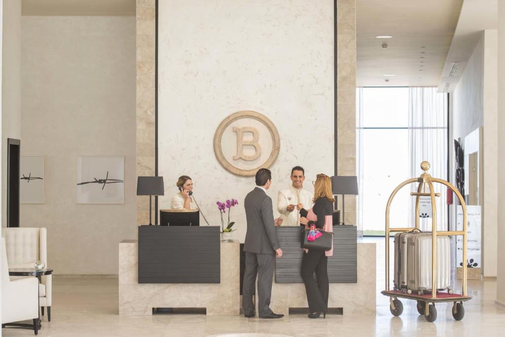 Luksusa viesnīcas reģistratūra ar viesmīlīgu personālu un apmeklētājiem, kas veic reģistrāciju. Uz fona redzams moderns interjers ar gaisīgu atmosfēru.
