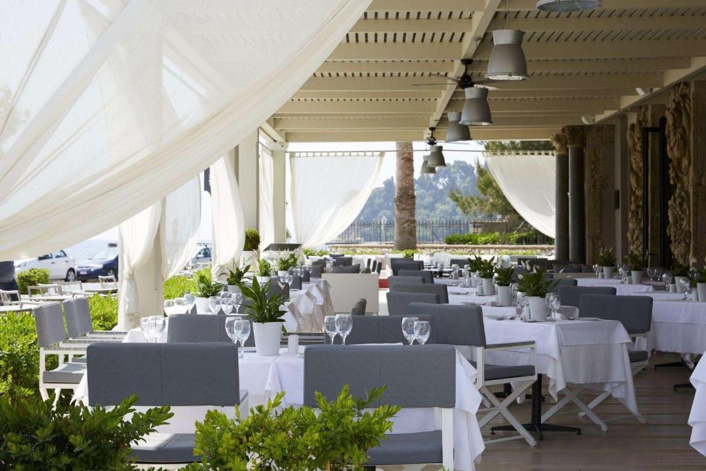 Elegants restorāns ar terasi, kurā ir sakārtotas baltas galdautu klātas galdi un zaļi augi. Gaisīgas aizkaru daļas rada patīkamu atmosfēru.