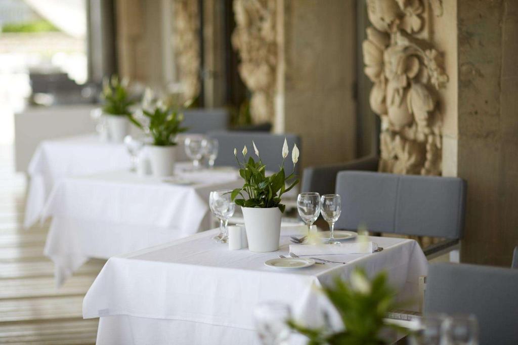 Elegants restorāna interjers ar baltiem galdautiem, zaļām augiem un glāzēm, kas rada patīkamu atmosfēru. Ideāli piemērots romantiskām vakariņām vai svinībām.