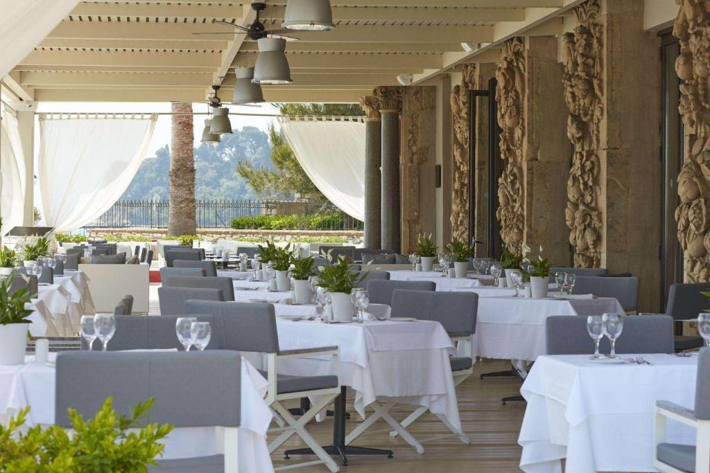 Elegants restorāns ar baltām galdautām un modernām mēbelēm, iekārtots ar zaļām augiem un ar skatu uz dabu. Gaisma plūst caur caurspīdīgām aizkariem, radot patīkamu atmosfēru.