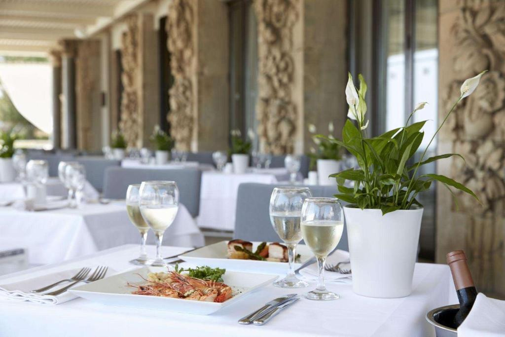 Elegants restorāna galds ar baltu galdautu, uz kura atrodas jūras veltes, vīna glāzes un dekoratīva puķe. Galds izvietots gaišā un stilīgā vidē, kas rada patīkamu atmosfēru.