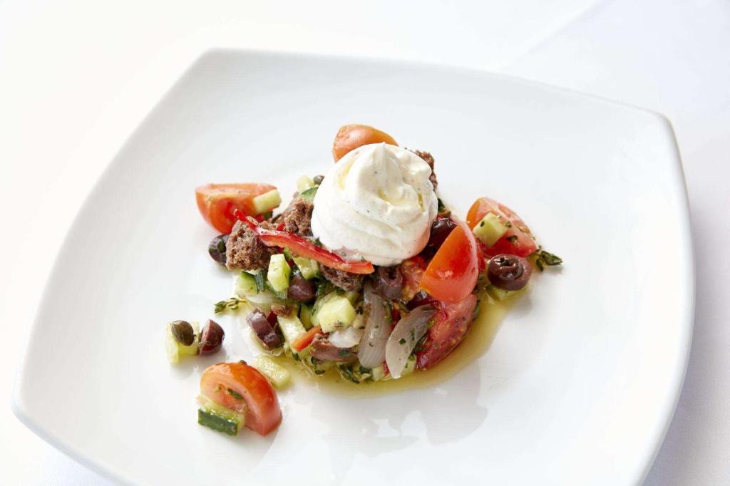 Svaiga dārzeņu salātu porcija ar tomātiem, gurķiem un melnajām olīvām, papildināta ar krēmīgu baltu mērci, pasniegta uz balta šķīvja.
