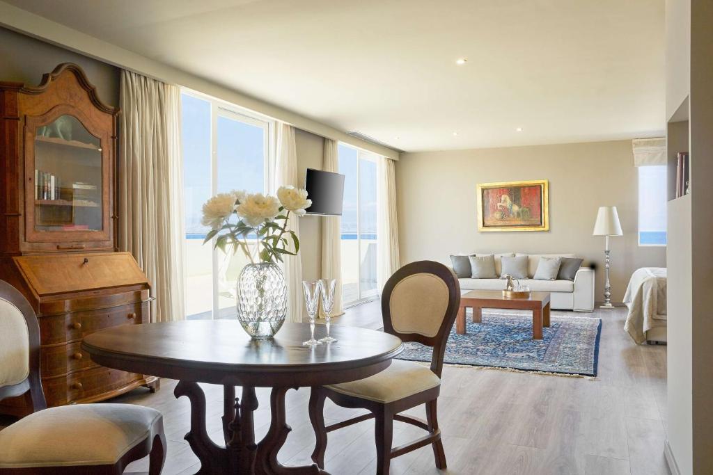 Mūsdienīgs viesistabas interjers ar koka mēbelēm, ziedu kompozīciju un plašiem logiem, caur kuriem redzams jūras skats. Eleganti izkārtota telpa ar dīvānu un mākslas darbu uz sienas.