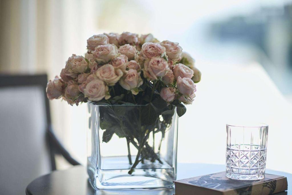Rožu pušķis caurspīdīgā vāzē, blakus kristāla glāze un grāmata uz galda, radot mierīgu un elegantu atmosfēru.