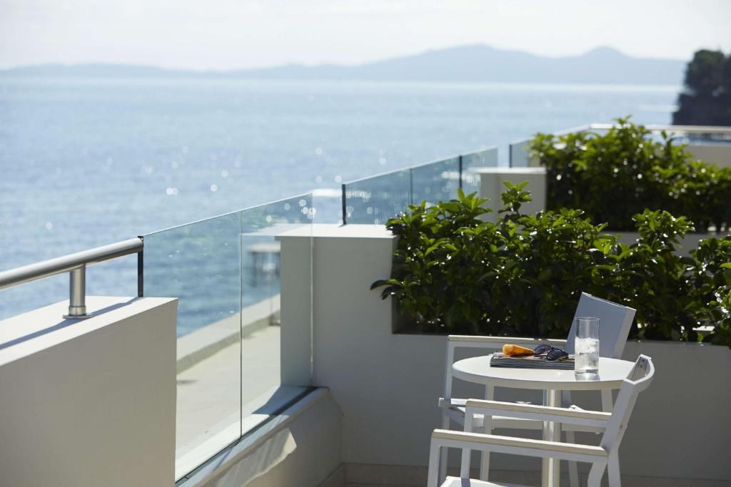 Terases skats uz jūru ar baltu galdu un krēsliem, uz kura atrodas glāze ūdens un saulesbrilles. Fons ar mīkstu, spožu ūdeni un kalnainu piekrasti.