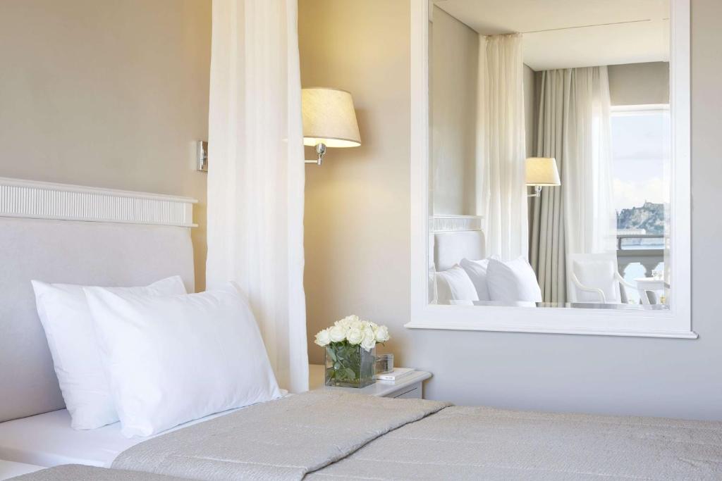 Mūsdienīgs viesnīcas numurs ar gaisīgu interjeru, kurā ir gulta, spilveni un ziedi uz naktsgaldiņa. Spogulī redzama telpa ar dīvānu un logu, caur kuru ieplūst dabiska gaisma.