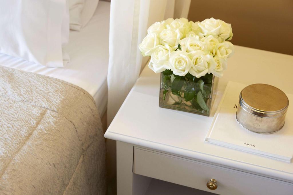 Balta rožu pušķis stikla traukā, novietots uz naktsgaldiņa blakus gultai, radot mājīgu un elegantu atmosfēru. Apakšā atrodas grāmata un sudraba krāsas trauks.
