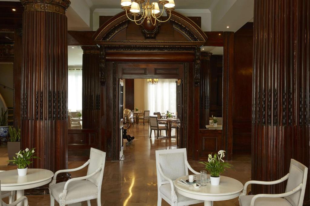 Eleganta interjera ar koka kolonnām un gaišiem logiem, kas ved uz ēdamzonu. Uz galda atrodas puķu pods un glāze ūdens.