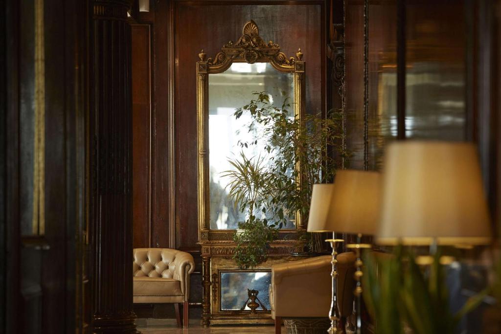 Elegantā viesnīcas interjera aina ar izsmalcinātu spoguli un dīvānu, papildināta ar zaļām augiem un gaismas ķermeņiem. Atmosfēra ir silta un aicinoša.