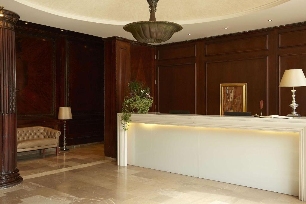 Elegants viesnīcas reģistratūras interjers ar tumšu koka apdari un mūsdienīgu pieņemšanas galdu. Telpā ir dīvāns un gaismas avoti, kas rada mājīgu atmosfēru.