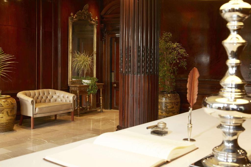 Elegantā viesnīcas uzgaidāmā telpa ar greznu dīvānu, spoguli un dekoratīvām augu kompozīcijām. Interjers izceļas ar siltu koka apdari un klasiskām mēbelēm.