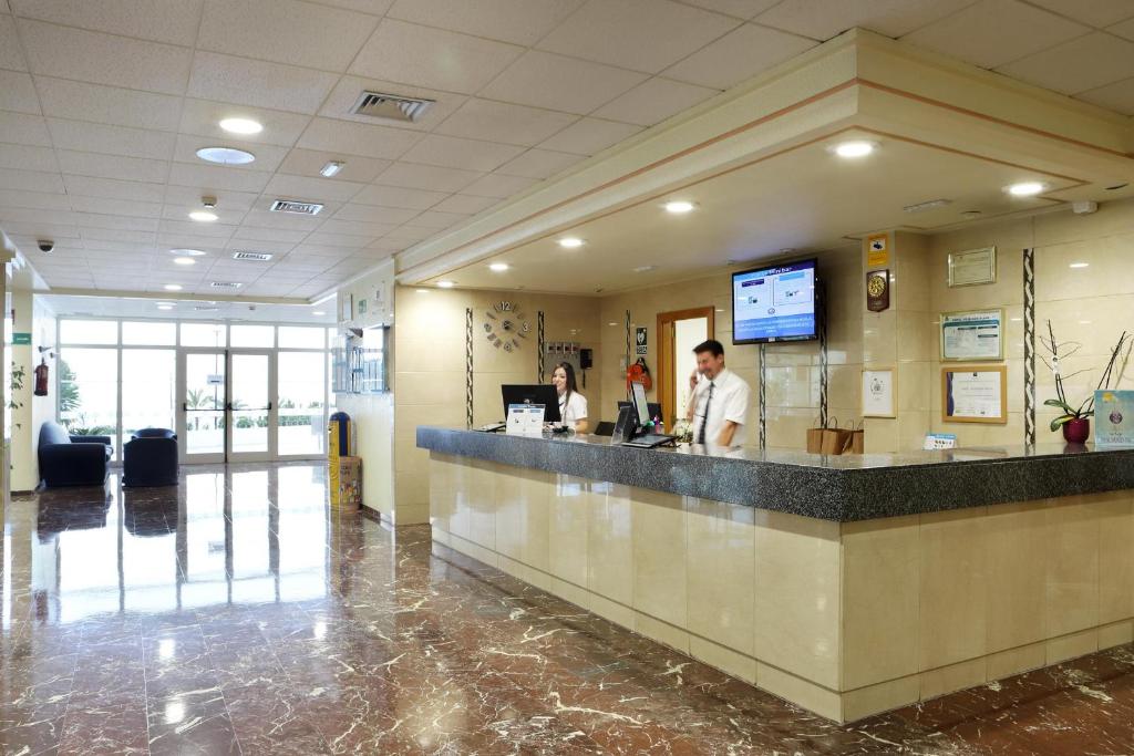 Mūsdienīgs viesnīcas reģistratūras interjers ar darbiniekiem, kas apkalpo viesus. Gaiša un plaša telpa ar marmora grīdu un lieliem logiem.