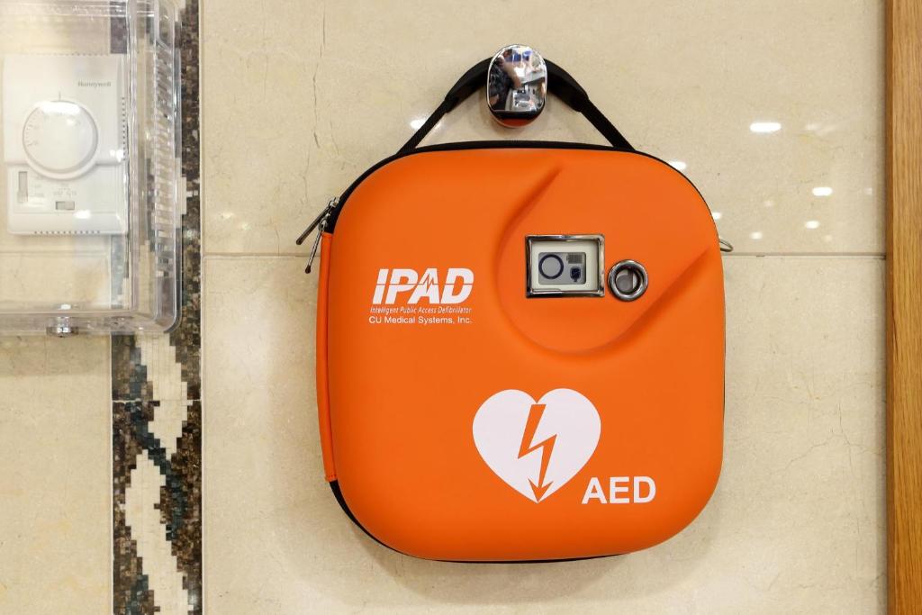 Oranžs sirds formas AED (automātiskais ārējais defibrilators) korpuss, kas uzstādīts uz sienas, ar skaidru zīmējumu un logu, lai redzētu ierīci iekšpusē.