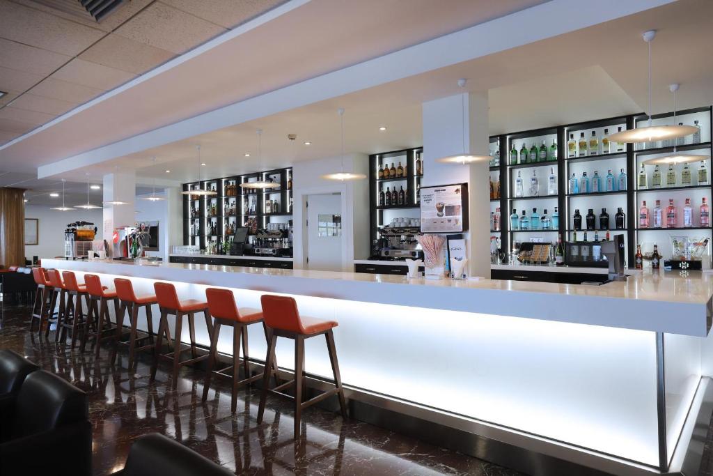 Mūsdienīgs bārs ar gaišu interjeru, izgaismoto letes daļu un stilīgām oranžām krēsliem, kas izkārtoti gar letes. Aiz letes ir redzami dažādu alkoholisko dzērienu plaukti, radot aicinošu atmosfēru.