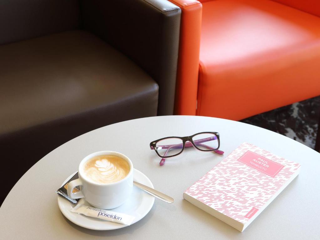 Kafijas tase ar sirds formu uzputotu virsotni, blakus glāze ar rozā un melnu rāmīti, un grāmata ar rozā vāku. Ideāla brīvdienu brīža attēls.