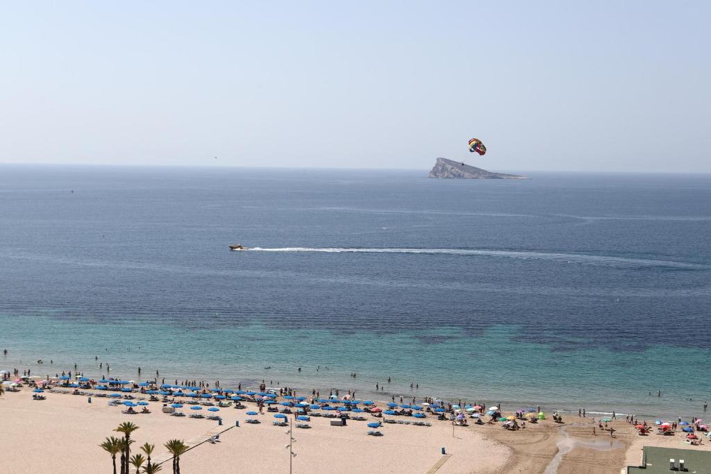 Pludmale ar saules lietām un cilvēkiem, kas izbauda brīvdienas, ar laivu un paraglidingu gaisā. Tālā horizonta daļā redzama sala.