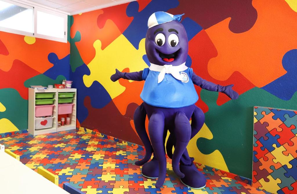 Mīksts purpursarkans astoņkājis tērpā, kas priecīgi pozē krāsainā bērnu telpā ar puzles formas grīdu un dzīvīgu sienu apdari.
