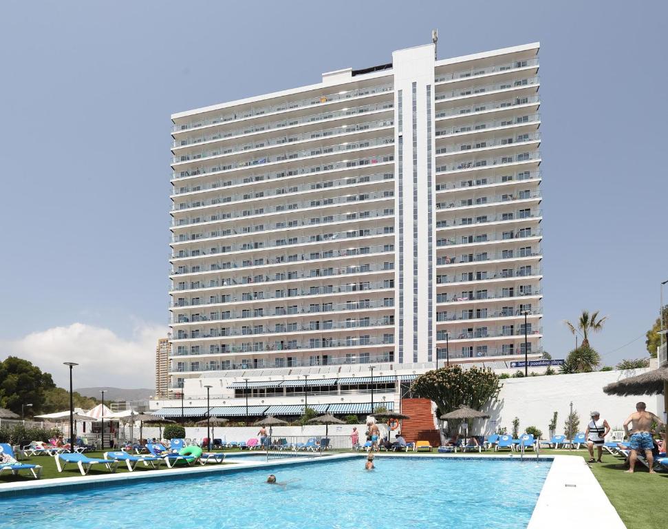 Liels viesnīcas komplekss ar baseinu un sauļošanās krēsliem, kas apņem to. Pilsētas ainava ar skaidru debesu fonu.