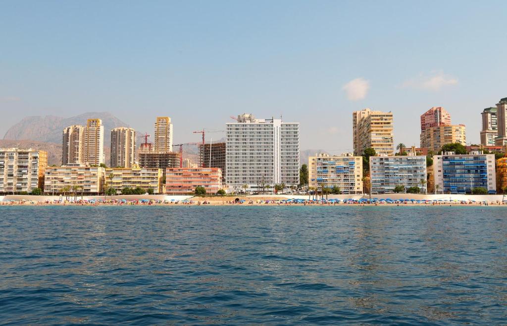 Pludmales skats uz pilsētu, kur redzami mūsdienīgi augstceltnieki un pludmale ar atpūtas vietām saulainā dienā.