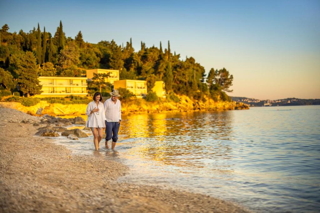 Pāris staigā pa pludmali pie saulrieta, baudot mierīgu brīdi ūdens malā, ar elegantām mājām kalnu fona.