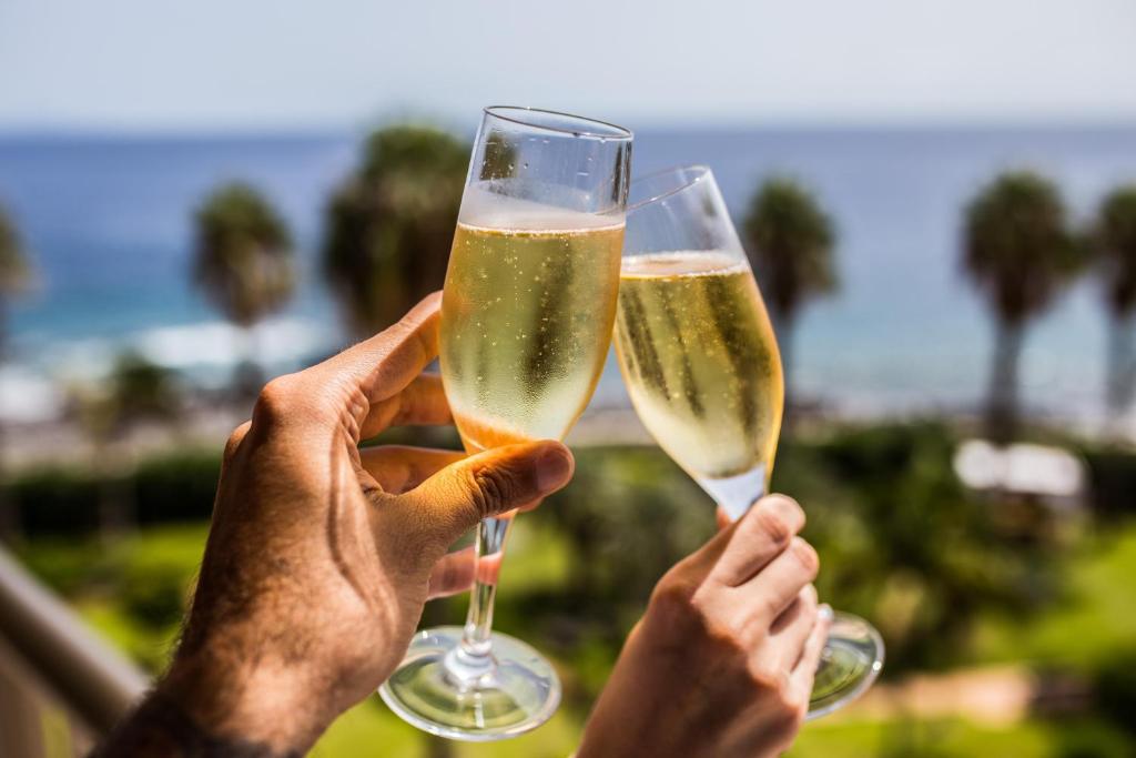 Divi cilvēki sveicina ar šampanieša glāzēm, fona redzams jūras skats un palmas. Ideāla brīvdienu atmosfēra.