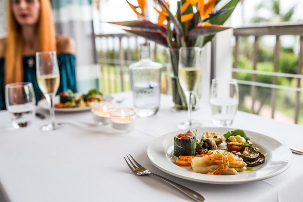 Elegants galda iestatījums ar gourmet ēdienu, dzērieniem un ziediem. Uz galda ir grezns ēdiens, kas sastāv no dārzeņiem un garnelēm, kā arī šampanietis un ūdens glāzes.