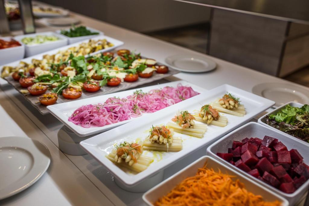 Krāšņa salātu bufete ar dažādām dārzeņu uzkodām, tostarp tomātiem, sīpoliem, gurķiem un burkāniem, kā arī skaisti pasniegtiem ēdieniem uz baltām šķīvēm.