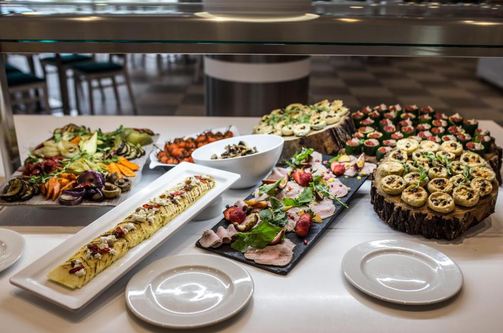 Izsmalcināts uzkodu galds ar dažādām delikatesēm, tostarp dārzeņu salātiem, gaļas šķēlītēm un konditorejas izstrādājumiem. Galds rotāts ar augļiem un radošiem pasniegšanas veidiem.